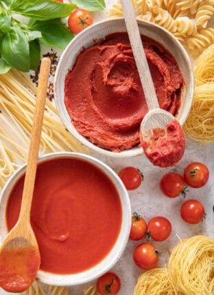 Two white bowls filled with tomato sauce and tomato paste. Dried pasta noodles are around the two bowls.