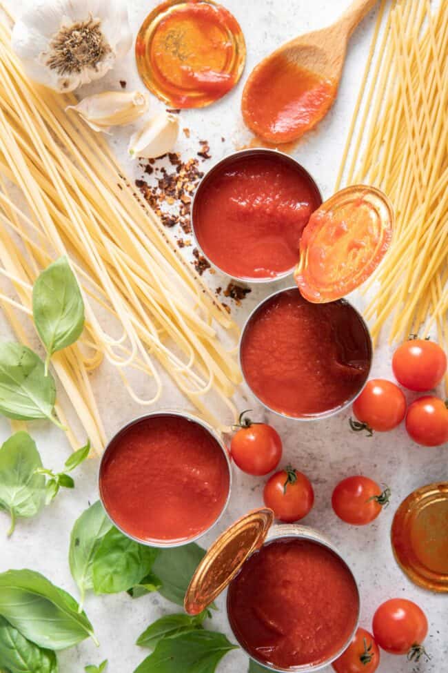 Four opened cans of red vegetable puree next to uncooked noodles and basil leaves.