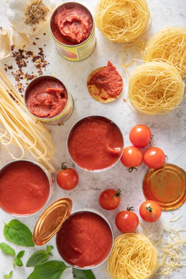 Two opened cans of tomato paste and three cans of opened tomato sauce. Dried noodles and fresh parsley sit next to the cans.