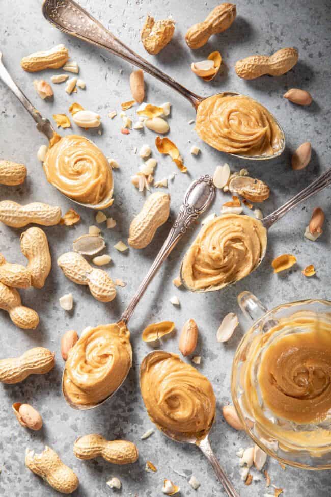 Five spoons filled with peanut butter. Peanuts and shells are scattered next to the spoons.