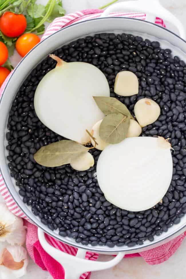 A white pot filled with dried black beans, a white onion sliced in half, bay leaves and garlic cloves.