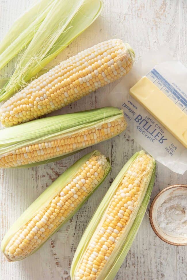 Four fresh corn on the cob set on a white wooden table. A stick of butter and a small dish of salt sit next to the corn.