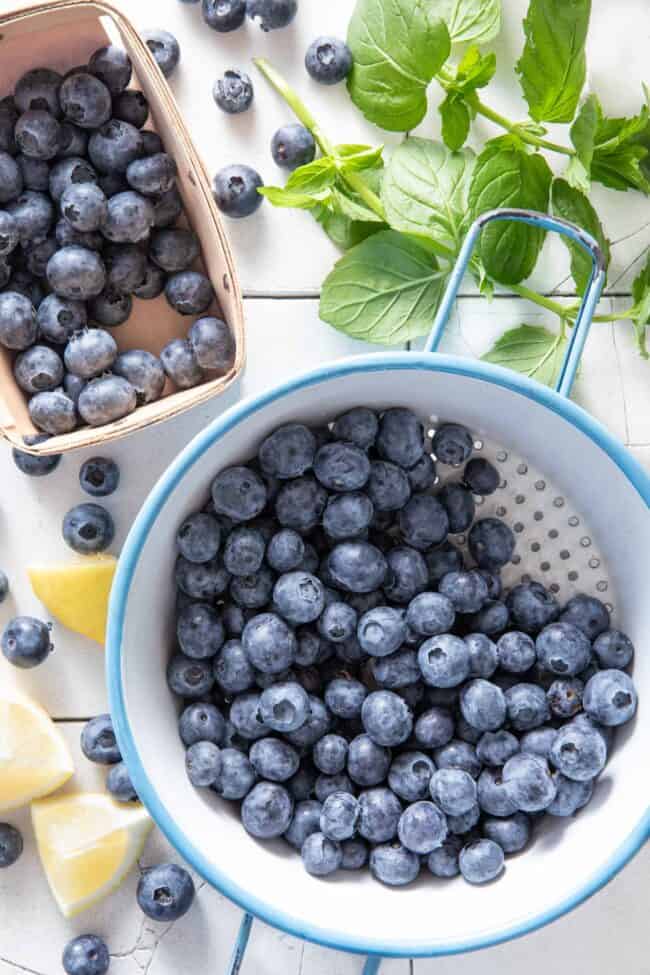 A blue and white colander filled with fresh blueberries. A box of blueberries sits next to the colander.