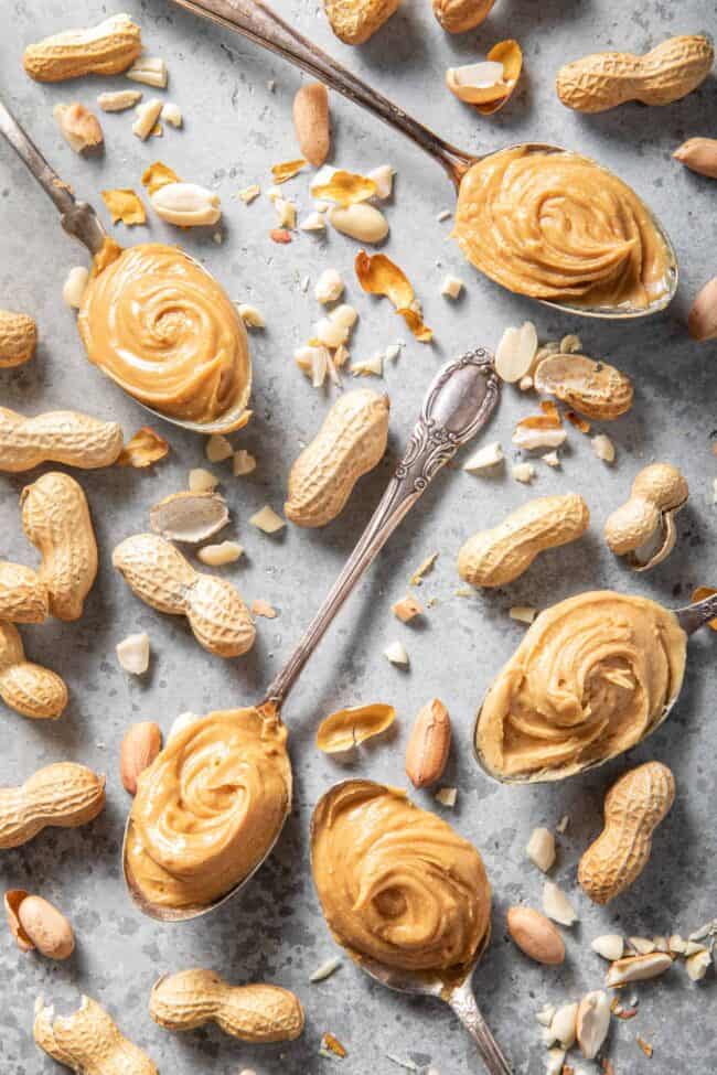 Five spoons filled with peanut butter. A clear glass cup has a swirl of peanut butter in it next to the spoons.