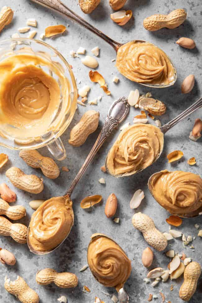Five spoons filled with peanut butter. A clear glass cup has a swirl of peanut butter in it next to the spoons. For how many tablespoons in 1/2 cup conversion.