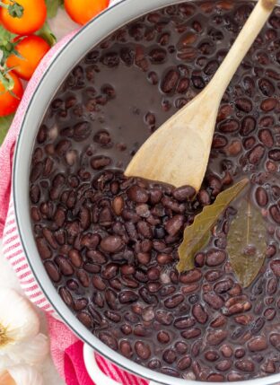 A white pot filled with cooked black beans. A wooden spoon rests inside the pot.