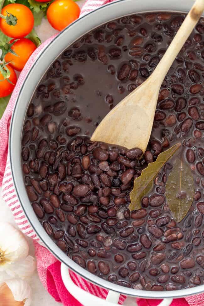 A white pot filled with cooked black beans. A wooden spoon is poked in the beans.