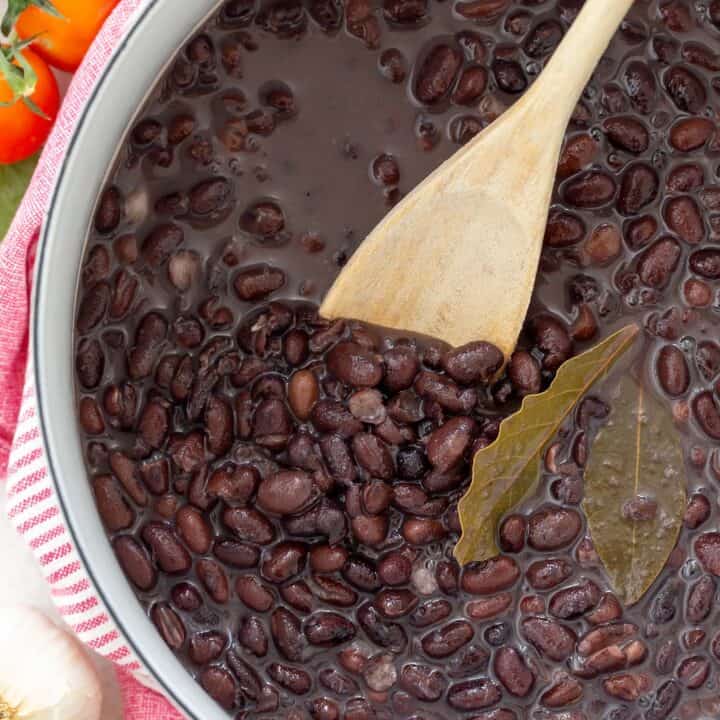 A white pot filled with cooked black beans. A wooden spoon rests inside the pot.