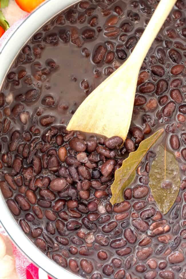 A white pot filled with cooked black beans from scratch. A wooden spoon rests inside the pot.