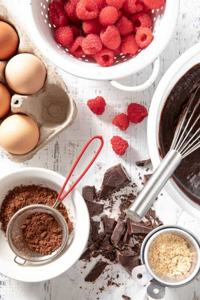 A carton of eggs sits next to a white mixing bowl filled with brownie batter. A bowl or raspberries sits next to them.