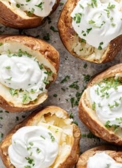 Six baked potatoes topped with sour cream and chives on a baking sheet.