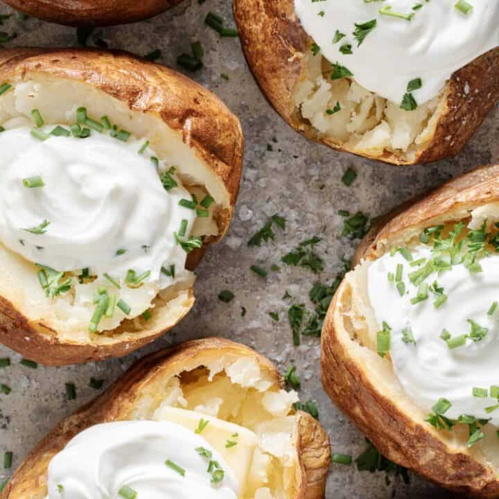 Six baked potatoes topped with sour cream and chives on a baking sheet.