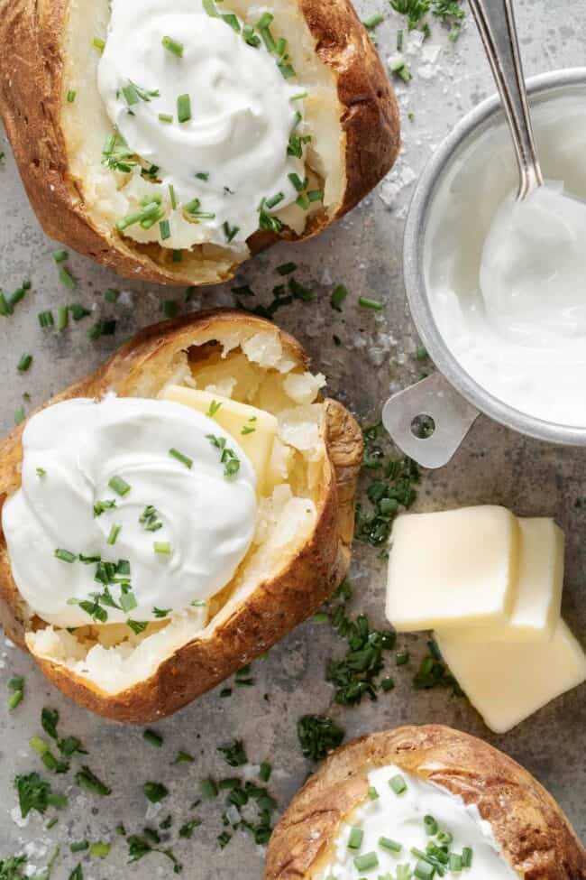 Three baked potatoes topped with sour cream. A aluminum measuring cup filled with sour cream with a spoon in it sits next to the baked potatoes.