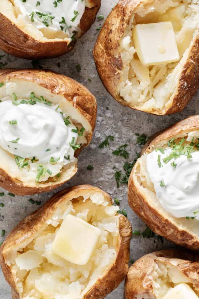 Six baked potatoes topped with butter and chives on a baking sheet.
