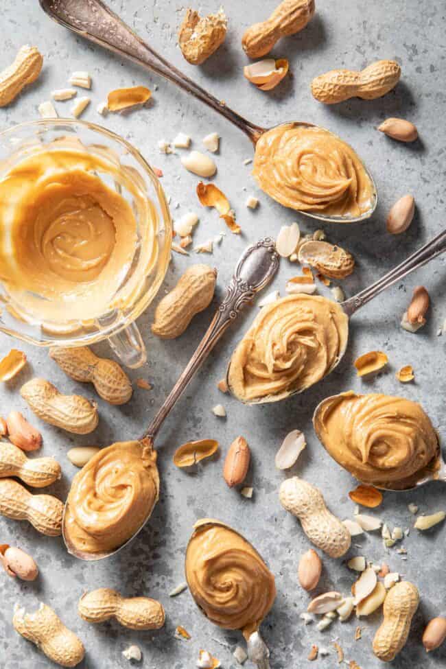 Five spoons filled with peanut butter. A clear glass cup has a swirl of peanut butter in it next to the spoons. For how many tablespoons in 1/2 cup conversion.