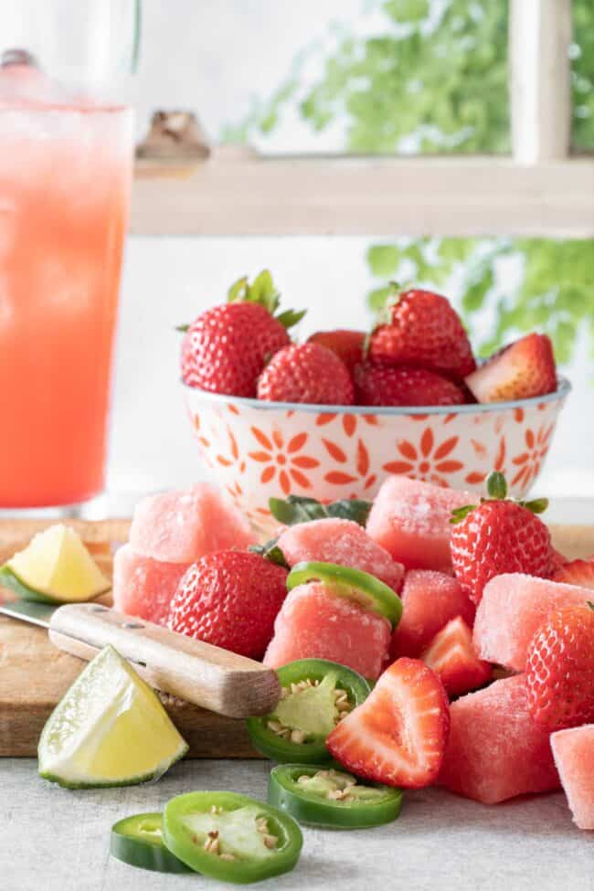 A red and white bowl filled with fresh strawberries sits on a wood cutting board. Next to the bowl sits a knife and sliced strawberries and jalapeno. A window with white window panes is behind the bowl of strawberries.