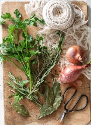 Fresh parsley, thyme and rosemary on a wood cutting board next to a ball of string and small scissors.