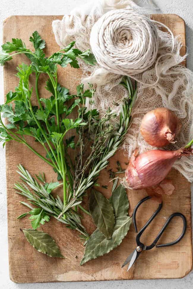 Fresh herbs, kitchen string and cheesecloth are set on a wood cutting board. 