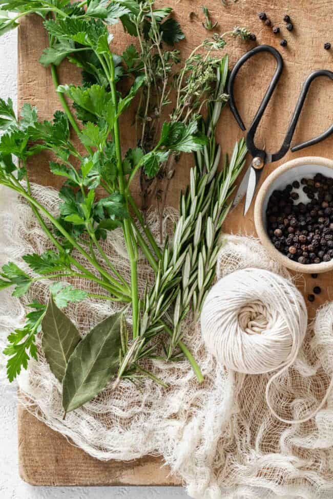 Fresh herbs, kitchen string and cheesecloth are set on a wood cutting board.