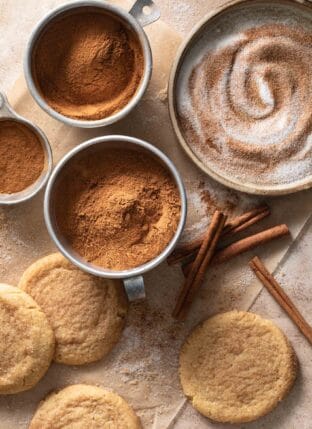 Aluminum measuring cups filled with cinnamon. Cinnamon sticks and snickerdoodle cookies sit next to the measuring cups.