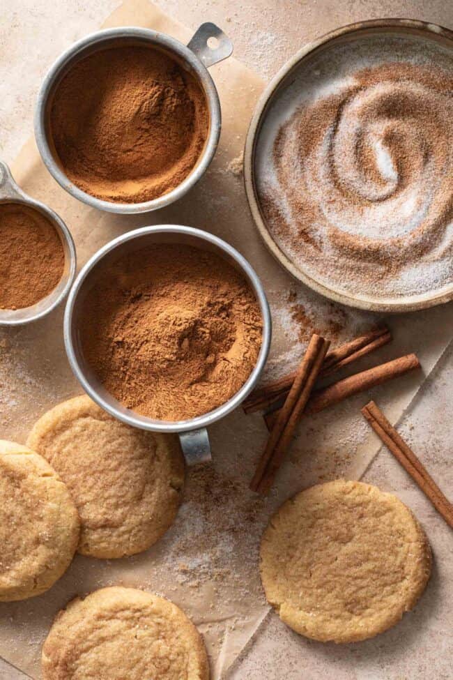 Aluminum measuring cups filled with cinnamon. Cinnamon sticks and snickerdoodle cookies sit next to the measuring cups - for cinnamon substitutes.