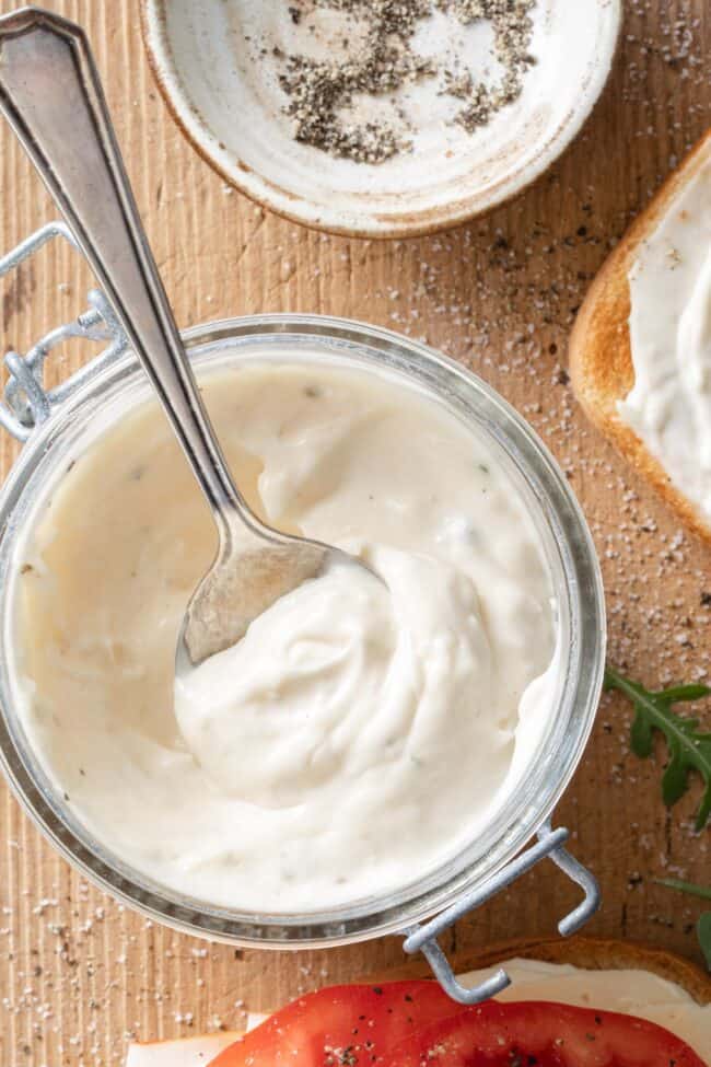 A clear glass jar filled with mayonnaise sauce. A spoon rests in the jar.