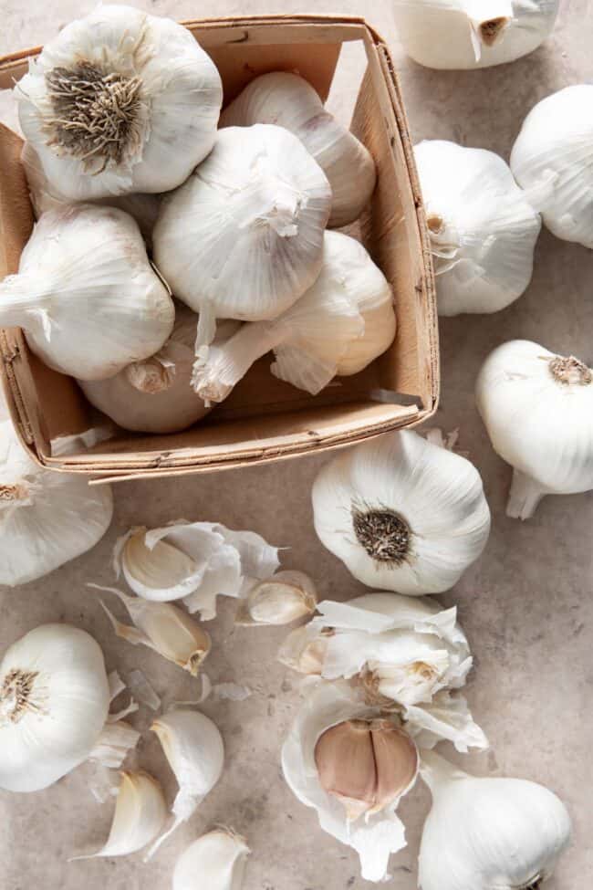 A small square basket filled with whole heads of garlic with cloves of garlic scattered next to the basket.