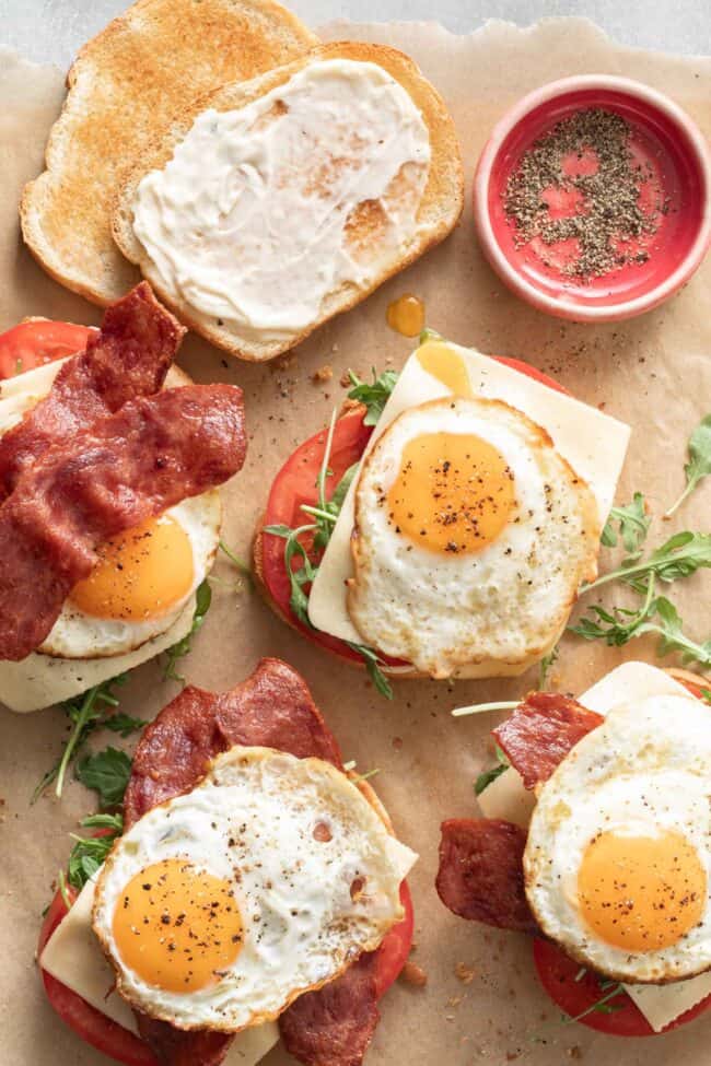 Four fried egg sandwiches with eggs, turkey bacon, cheese, arugula and tomato sit on a piece of parchment paper.