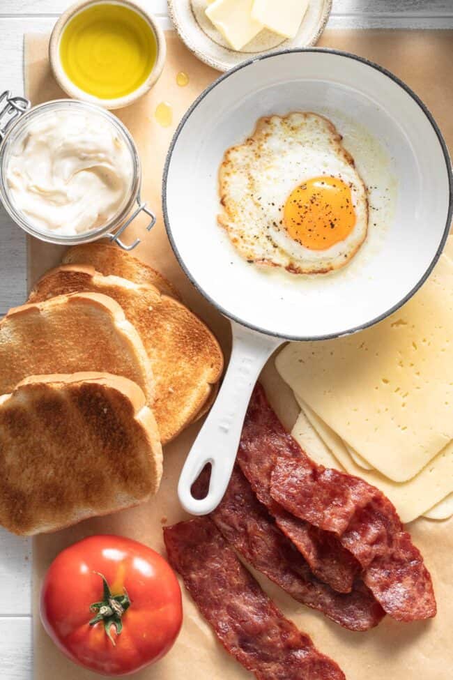 Pieces of toast sit next to a tomato, cheese slices, cooked bacon and a glass jar filled with mayonnaise.