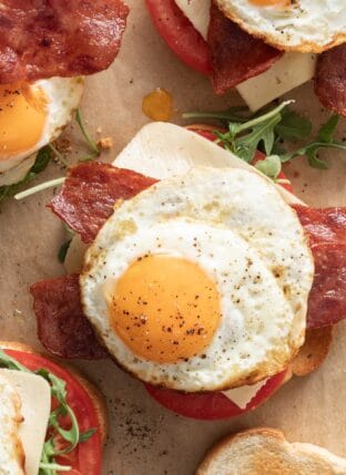 A fried egg sandwich with eggs, turkey bacon, cheese, arugula and tomato on a piece of parchment paper.