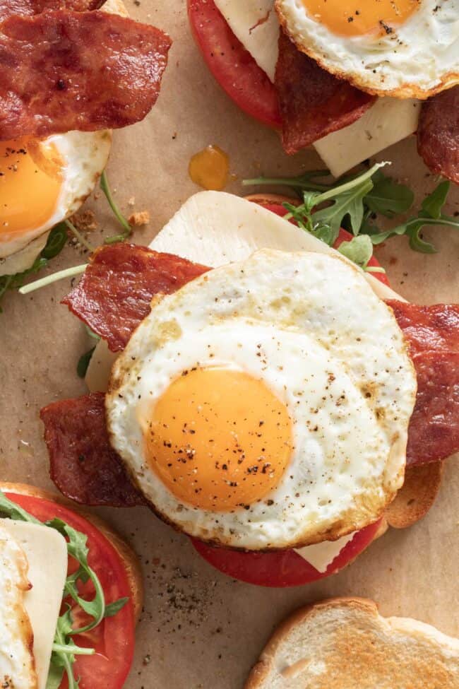 A fried egg sandwich with eggs, turkey bacon, cheese, arugula and tomato on a piece of parchment paper.