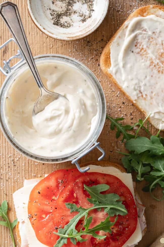 A sandwich on a wooden cutting board next to a small glass jar filled with garlic aioli (mayonnaise with garlic).