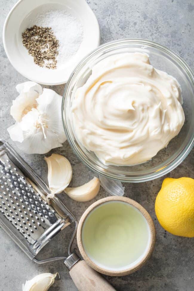 A small clear glass mixing bowl filled with mayonnaise, next to a head of garlic, lemon and a small bowl of lemon juice.