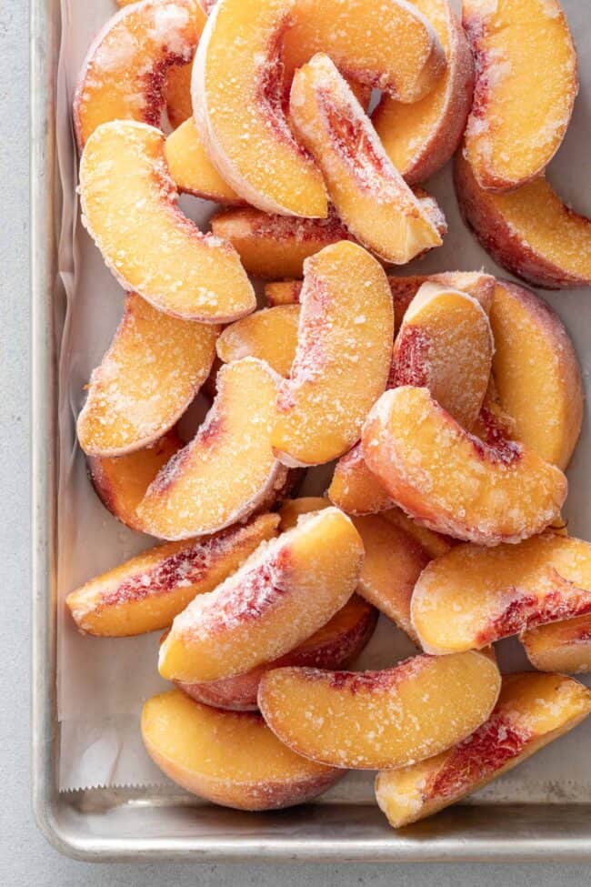 Many frozen peach slices piled on a cookie sheet.