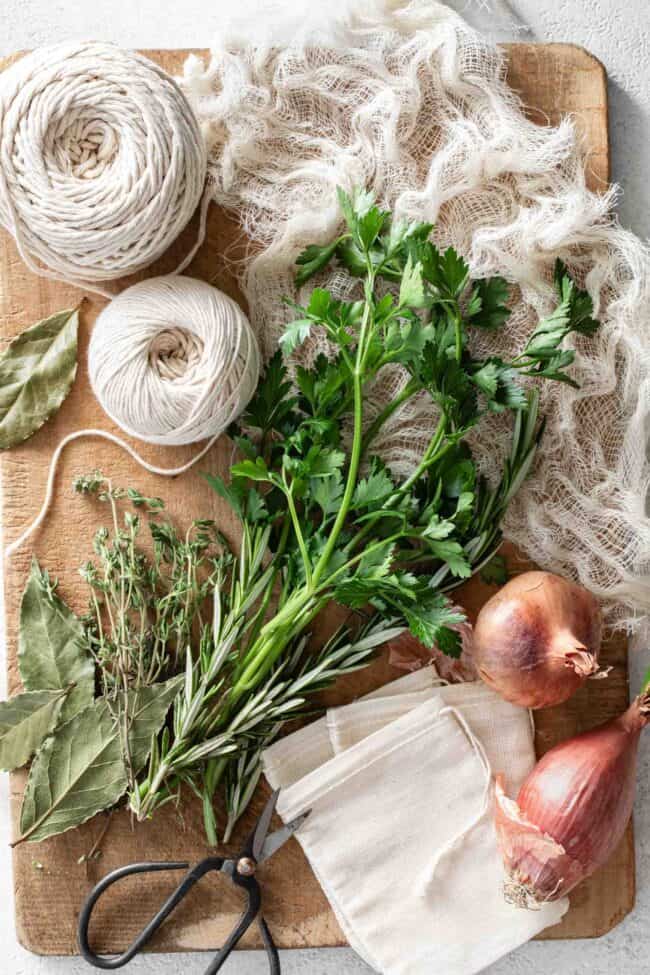Fresh herbs, kitchen string and cheesecloth to make a bouquet garni are set on a wood cutting board.
