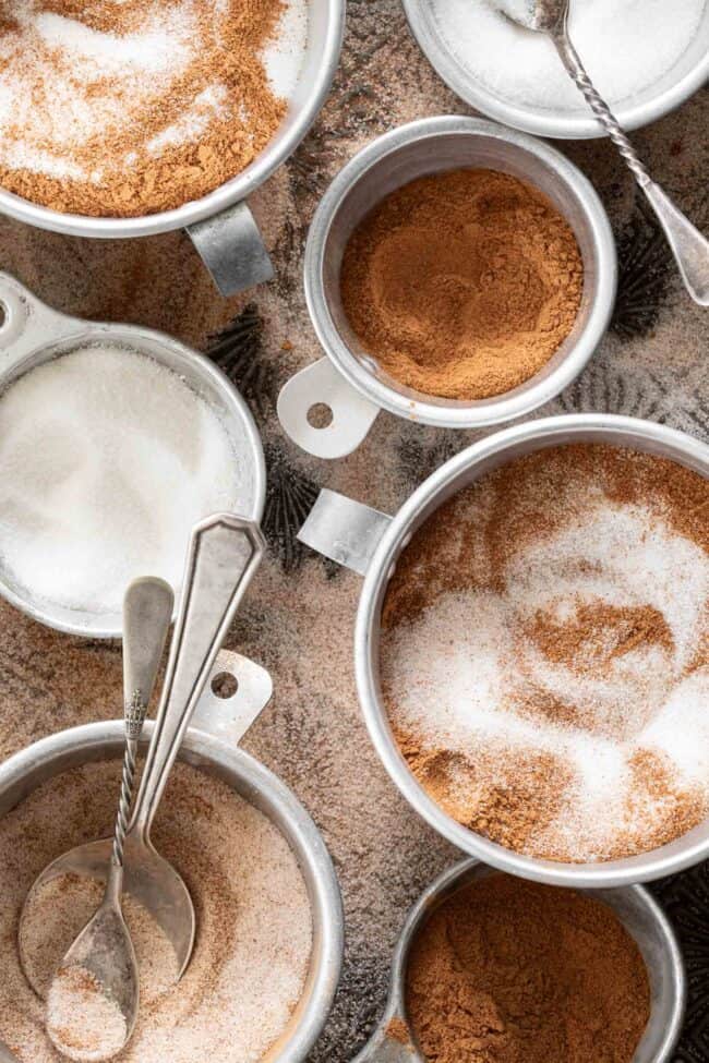 Aluminum measuring cups filled with cinnamon, sugar and some mixed with both cinnamon sugar.
