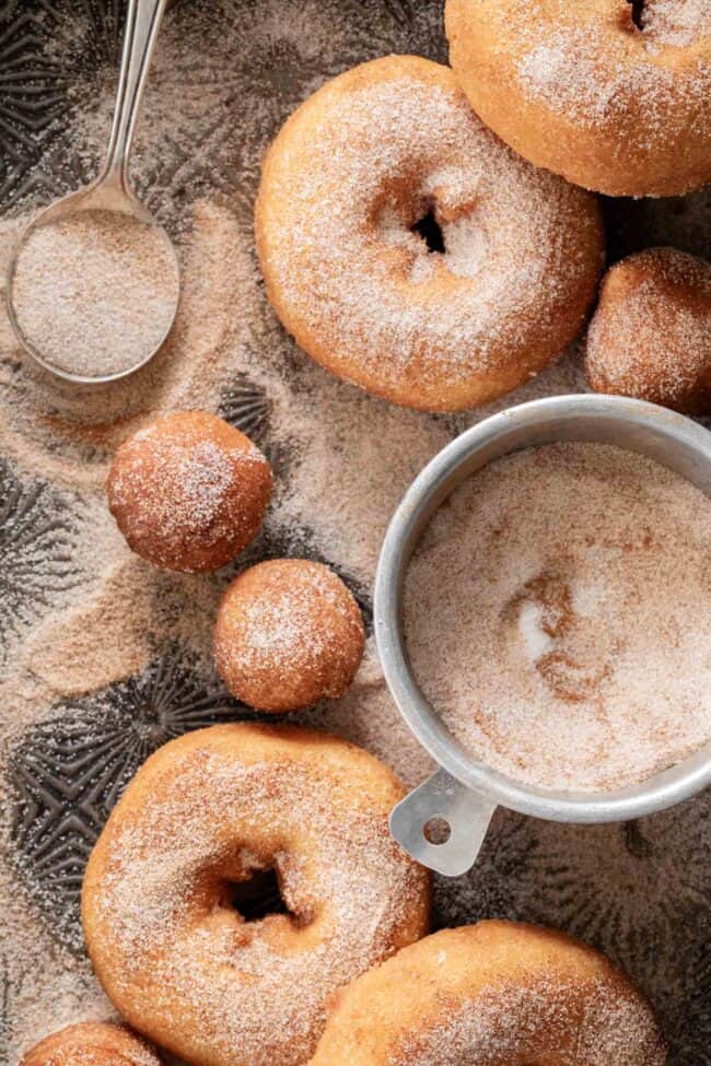 Plain donuts dusted with cinnamon sugar on a cookie sheet with a spoon filled with cinnamon sugar.
