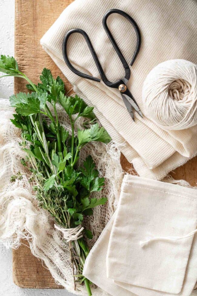 A bundle of fresh herbs (bouquet garni) are tied together and sit on a wood cutting board with cheesecloth, string, sachet bags and scissors.