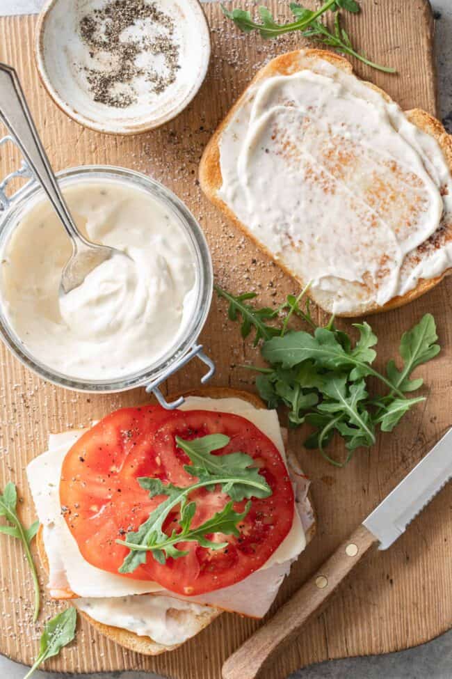 A sandwich on a wooden cutting board next to a small glass jar filled with garlic aioli (mayonnaise with garlic).