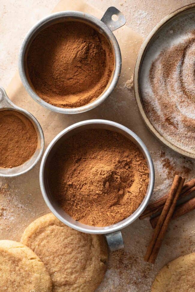 Aluminum measuring cups filled with cinnamon. Cinnamon sticks and snickerdoodle cookies sit next to the measuring cups - for cinnamon substitutes.