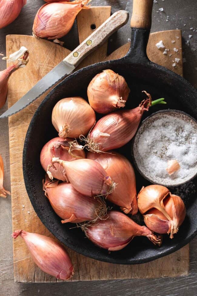 Several shallots sit in a cast iron pan next to a knife for shallot substitutes.