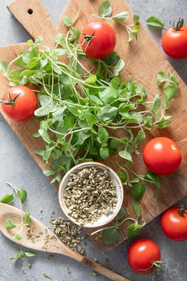 Fresh oregano and a small dish of dried oregano sit on a wood cutting board next to cherry tomatoes and a wooden spoon.
