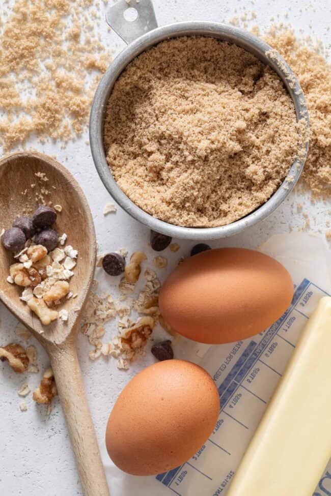 An aluminum measuring cup filled with brown sugar sits next to two eggs and a stick of butter.