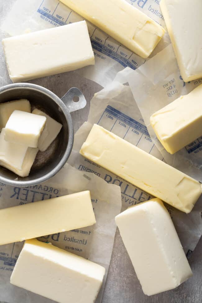 Several sticks of butter and wrappers sit next to an aluminum measuring cup filled with slices of butter. (For how many sticks of butter is 1 cup)