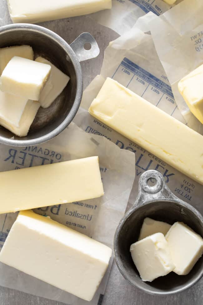 Several sticks of butter and wrappers sit next to an aluminum measuring cup filled with slices of butter.