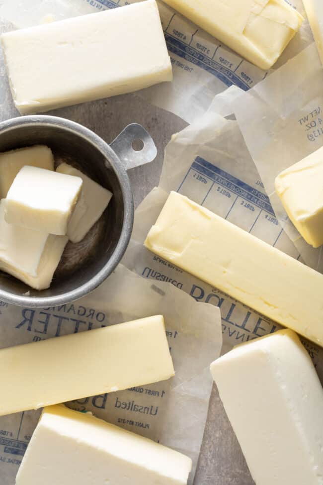 Several sticks of butter and wrappers sit next to an aluminum measuring cup filled with slices of butter.