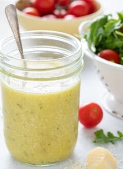 A mason jar filled with Parmesan Vinaigrette sits next to a white colander filled with salad greens.