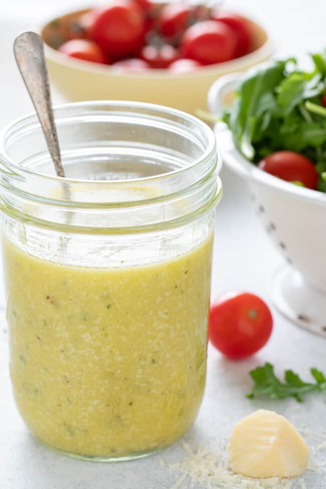 A mason jar filled with lemon dressing sits next to a white colander filled with mixed green lettuce.