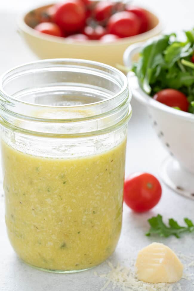 A mason jar filled with lemon dressing sits next to a white colander filled with mixed green lettuce.