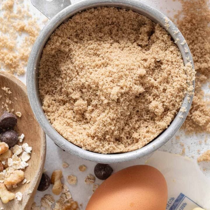 An aluminum measuring cup filled with brown sugar sits next to two eggs and a stick of butter.